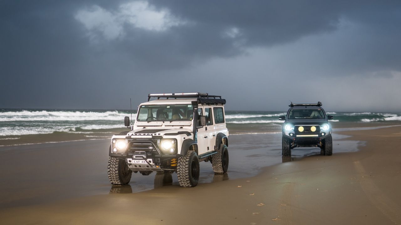 Two Land Rover Defenders with headlights on, on a beach under a storm