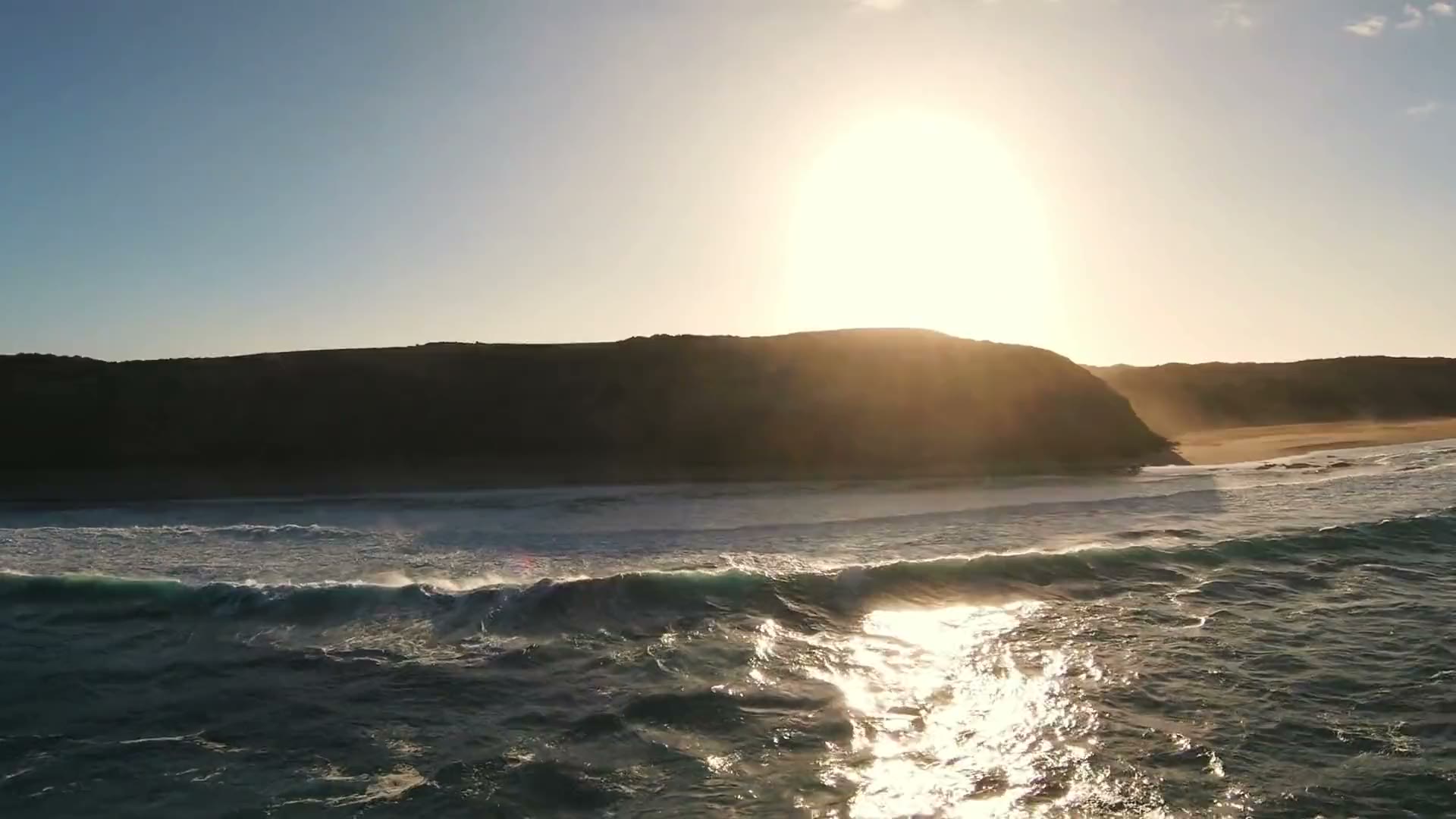 A headland in silhouette against a low sun, waves breaking below, from the air